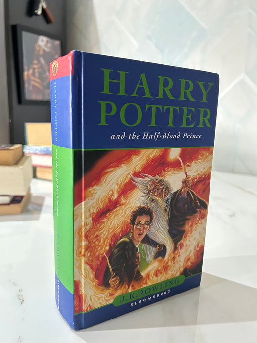 A hardcover copy of the Harry Potter book, 'Harry Potter and the Half-Blood Prince', standing upright on a white marble-like surface. The book features a blue spine and front cover with green title text and a colorful illustration of characters surrounded by flames.